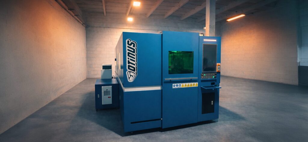 Enclosed industrial Otinus CNC laser cutting machine in a blue housing, installed in a production hall and equipped with a control panel and a viewing window, ensuring safe and precise metal processing.